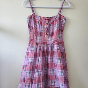 Peut inclure: Une robe sans manches avec un motif à carreaux rose, rouge et blanc. Le corsage présente une encolure carrée avec de fines bretelles et une patte de boutonnage ornée de quatre petits boutons rouges en forme de fraise. La jupe est froncée.
