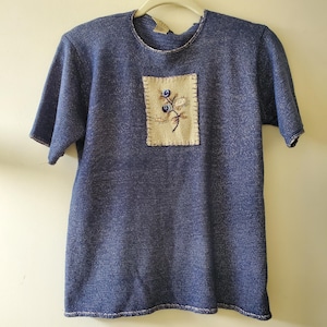 Puede incluir: Una camiseta de punto azul con mangas cortas y un parche cuadrado beige bordado con flores. El parche presenta bordados florales azules y marrones. La camiseta tiene un cuello redondo y un acabado texturizado.