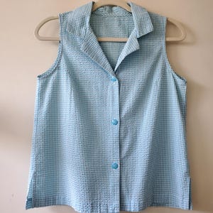 May include: A sleeveless, light blue and white checkered button-down shirt. The shirt has a collared neckline and is hanging on a beige hanger. The fabric has a textured, seersucker appearance.