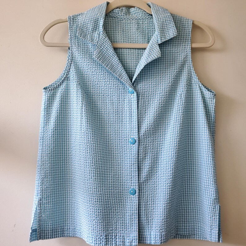 May include: A sleeveless, light blue and white checkered button-down shirt. The shirt has a collared neckline and is hanging on a beige hanger. The fabric has a textured, seersucker appearance.