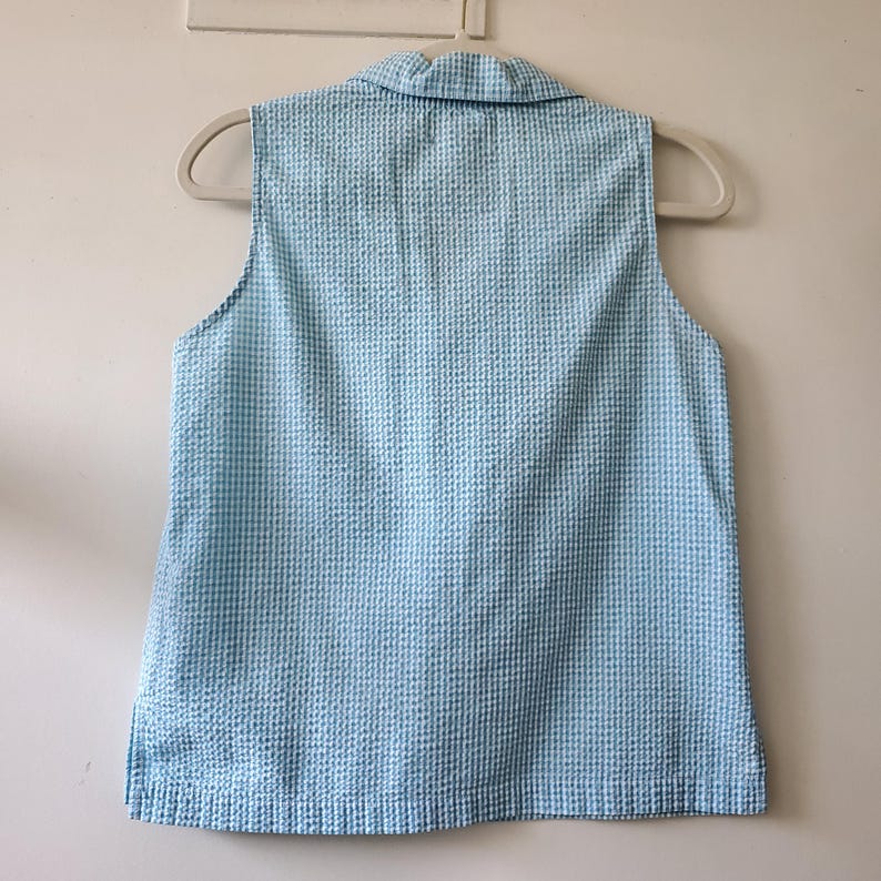 May include: A sleeveless, light blue seersucker top with a collared neckline. The fabric has a textured, checkered pattern. The garment is hanging on a white hanger against a plain white background.