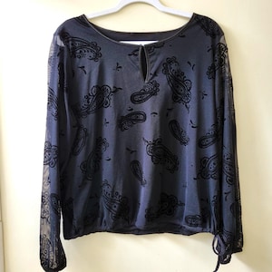 May include: A black long-sleeve top featuring a velvet-textured paisley pattern across the front and sheer mesh sleeves. The garment has a round neckline with a small vertical keyhole cutout and a gathered hemline with a tie detail at the side.