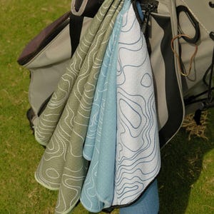 May include: A golf bag with several golf towels hanging from it. The towels are various shades of green and blue, with a white topographic map design. The golf bag is gray and black, and the background is green grass.