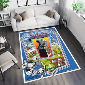 May include: A blue and white rug with a retro video game design. The rug features the Atari logo and the text "XEVIOUS". The rug also features a spaceship and a video game arcade cabinet.
