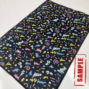 May include: Black rug with a geometric pattern in pink, blue, yellow, and white. The pattern includes circles, lines, and squiggles. The rug has the word "SAMPLE" printed on it in red.