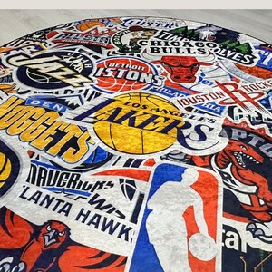 May include: A colourful rug featuring a variety of NBA team logos, including the Lakers, Nuggets, Bulls, Pistons, and Hawks. The rug is made from a soft, plush material and features a range of colours, including red, blue, yellow, and white.