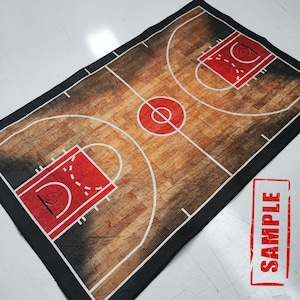 May include: A black and white basketball court design printed on a fabric rug. The rug features a brown wood grain pattern and white lines marking the court. The rug has a black border.