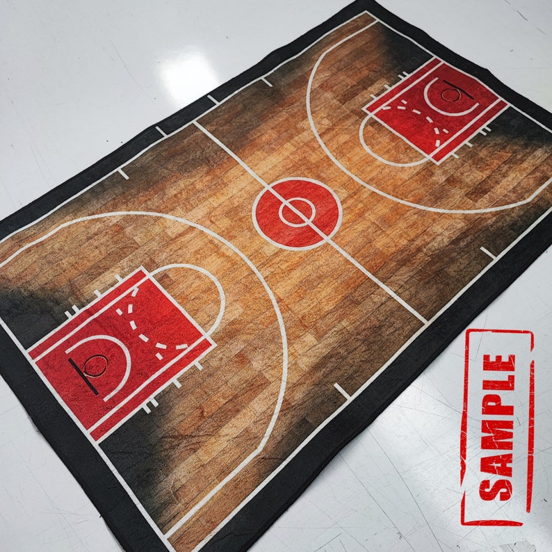 May include: A black and white basketball court design printed on a fabric rug. The rug features a brown wood grain pattern and white lines marking the court. The rug has a black border.