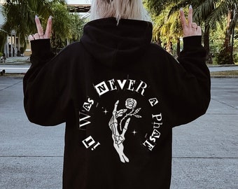 May include: Black hoodie with white graphic on the back. The graphic includes the text "I WAS NEVER A PHASE" and a skeleton hand holding a rose. The person is wearing white sneakers.