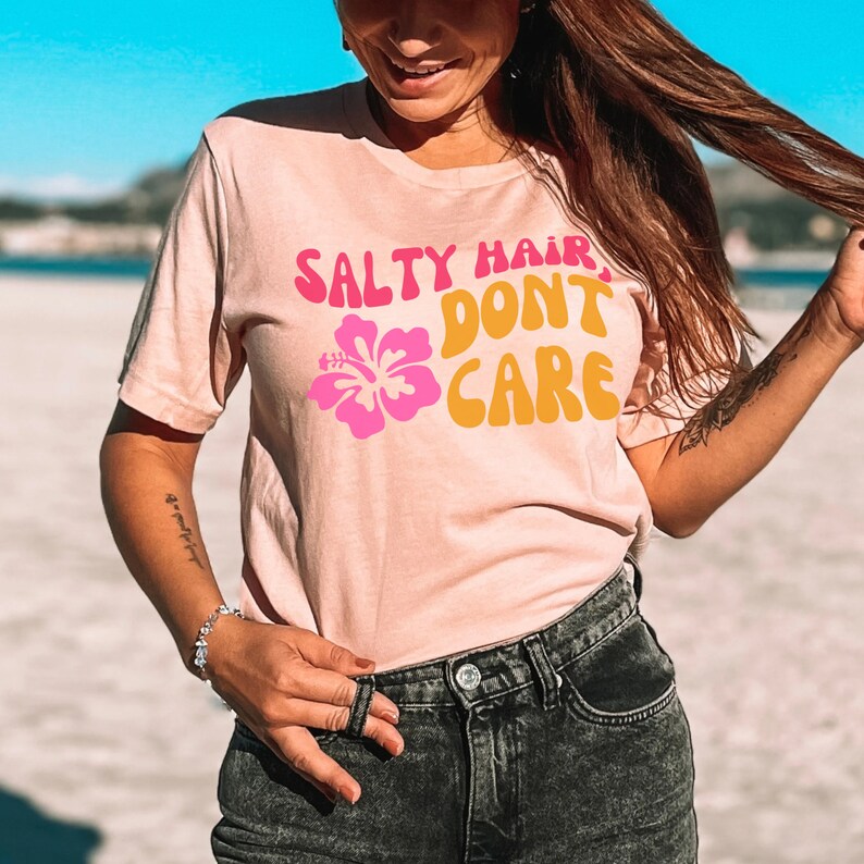 Peut inclure: Un t-shirt rose avec le texte "Salty Hair Don't Care" dans une police r&eacute;tro. Une fleur d'hibiscus rose est imprim&eacute;e sur le t-shirt. Le mannequin porte un short en jean noir.
