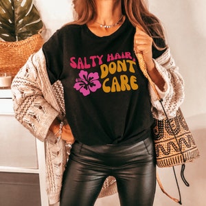 Peut inclure: Un t-shirt noir avec le texte "Salty Hair, Don't Care" dans une police r&eacute;tro. Une fleur d'hibiscus rose est &agrave; gauche du texte. Le mannequin porte le t-shirt avec une veste en cuir marron et un pantalon en cuir noir.