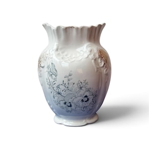 May include: A white ceramic vase with a scalloped rim and a bulbous body. The surface features a blue floral transfer print of pansies on the front, complemented by embossed scrollwork details and subtle gold-toned accents on the upper sides.
