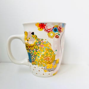 May include: White ceramic mug featuring a colorful cat design. The cat is adorned with yellow, blue, and floral patterns. The mug has a curved handle and is decorated with flowers and polka dots.