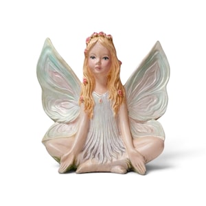 May include: A figurine of a seated fairy with long blonde hair adorned with small pink flowers. The fairy wears a white dress and features large, translucent wings with soft green and pink hues. The figure is posed with legs crossed.