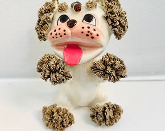 May include: A white ceramic poodle figurine with brown fur made of yarn. The poodle has a pink tongue sticking out and brown eyes.