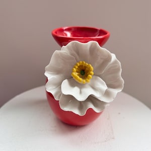 May include: A red ceramic oil burner featuring a large, three-dimensional white flower with a yellow center attached to the front. The top of the burner has a circular, bowl-shaped indentation for holding oil or wax, finished in a glossy red glaze.