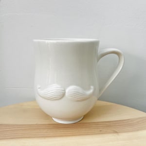 May include: A cream-colored ceramic mug with a sculpted mustache design. The mug has a curved handle and sits on a wooden surface. The mustache is a prominent feature, adding a touch of whimsy to the design. The mug is suitable for hot or cold beverages.