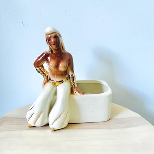 May include: A vintage ceramic planter featuring a seated figure with tan skin, blonde hair, and gold accents on the arms and waist. The figure is wearing white pants with gold trim. The planter is a rectangular shape and is cream-colored.