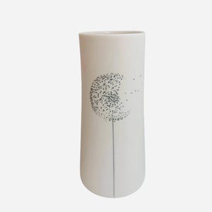 May include: A cylindrical white vase featuring a black line drawing of a dandelion. The design shows a single thin stem extending upward, topped with a circular seed head that appears to be dispersing small black dots toward the right side.