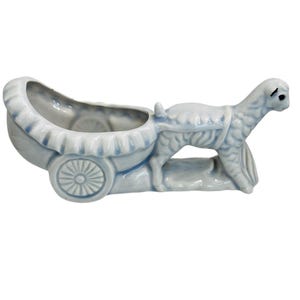 May include: A light blue ceramic planter shaped like a carriage pulled by a lamb. The carriage has a ruffled edge and a spoked wheel. The lamb is detailed with textured fur and stands on a small base. A decorative piece.