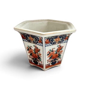 May include: A hexagonal ceramic planter featuring a white base with intricate floral patterns in orange, blue, and green. The rim is accented with a thin gold-colored line, and the exterior panels display detailed botanical illustrations.
