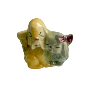 May include: A ceramic figurine of two sleeping dogs. One dog is yellow, the other is light green with a dark red bow. The dogs are nestled together, creating a charming and sentimental decorative piece.