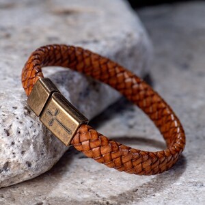 May include: A brown leather bracelet with a woven design and a metal clasp featuring a cross.