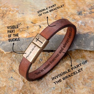 May include: A brown leather bracelet with a gold clasp. The clasp has a cross and the text "PSALMS 34:18". The bracelet is engraved with the words "You're mine forever and always papi 20".