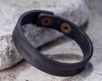 May include: Black leather bracelet with the text "BE LOVING, STRONG AND KIND - YOU" engraved on it.