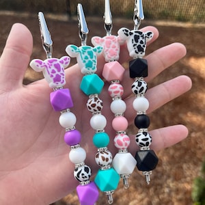 May include: Four beaded pacifier clips with cow-shaped charms. Each clip features a different color scheme: purple, teal, pink, and black. The beads are various shapes and sizes, including geometric and round, with silver accents.