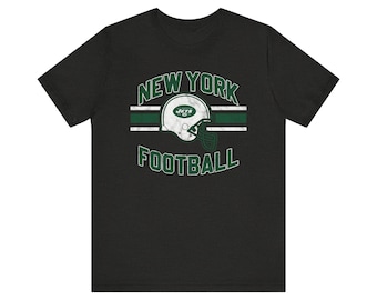 以下が含まれることがあります： 黒いTシャツ。緑と白で「NEW YORK FOOTBALL」の文字とフットボールのヘルメットのグラフィック。デザインには、中央に緑と白のストライプバンドが含まれています。