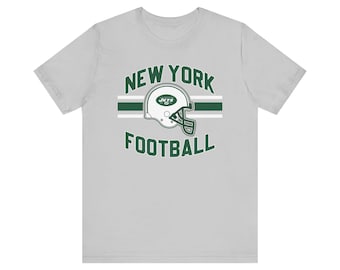 以下が含まれることがあります： 「NEW YORK FOOTBALL」の緑色の文字と、白と緑のアメリカンフットボールのヘルメットのグラフィックが特徴のライトグレーのTシャツ。クルーネックと半袖です。