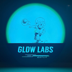 Puede incluir: Un disco que brilla en la oscuridad con un personaje de dibujos animados de un cr&aacute;neo lanzando un globo ocular. El disco tiene el texto "Glow Labs" y "Get Lit" impreso en &eacute;l.