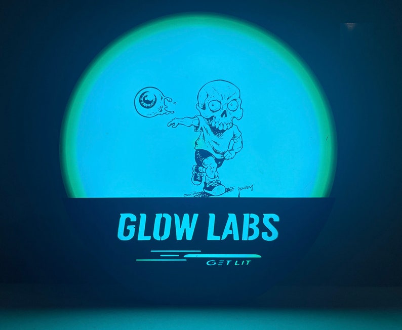 Puede incluir: Un disco que brilla en la oscuridad con un personaje de dibujos animados de un cr&aacute;neo lanzando un globo ocular. El disco tiene el texto "Glow Labs" y "Get Lit" impreso en &eacute;l.