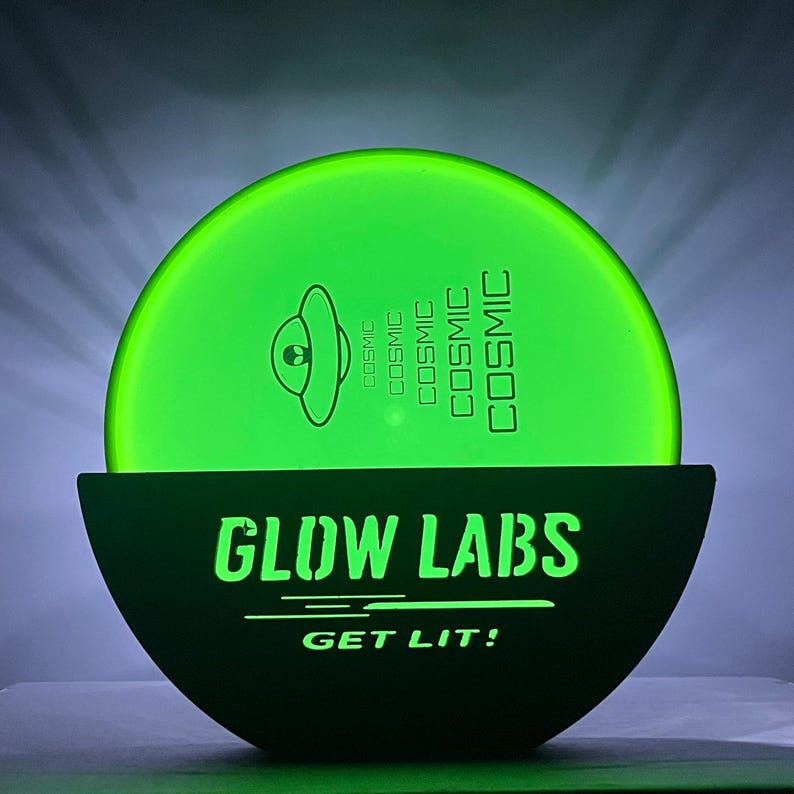 Puede incluir: Un disco verde que brilla en la oscuridad con un OVNI y el texto "COSMIC" impreso en &eacute;l. El disco est&aacute; sentado sobre una base negra con el texto "GLOW LABS GET LIT!" impreso en &eacute;l.