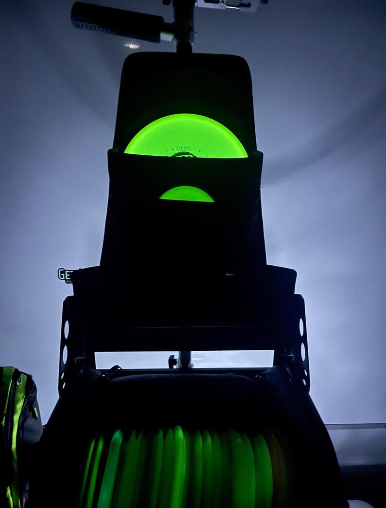 Puede incluir: Una bolsa negra de disco de golf con un disco verde que brilla en la oscuridad en el bolsillo superior. La bolsa tiene varios bolsillos y una correa. La bolsa est&aacute; llena de m&aacute;s discos que brillan en la oscuridad.
