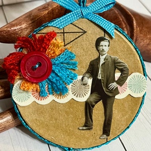 May include: A round, brown paper ornament with a blue ribbon, a red button, and a vintage photo of a man in a suit. The ornament is decorated with colorful fabric scraps and a geometric design.