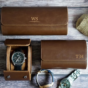 May include: Three brown leather watch cases with snap closures. One case is open, revealing a black chronograph watch with a silver bezel. The other two cases are closed and feature gold-stamped monograms reading WS and T|H on the front flaps.