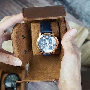 May include: A brown leather watch winder with a blue watch with a rose gold face and hands. The watch has a chronograph and the words "Fossil" on the face.