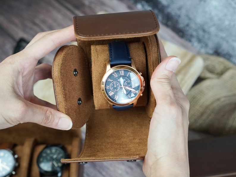 May include: A brown leather watch winder with a blue watch with a rose gold face and hands. The watch has a chronograph and the words "Fossil" on the face.
