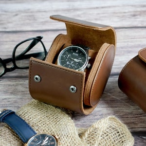 May include: A brown leather watch roll case with a snap button closure containing a black Boss wristwatch with a silver bezel. A second wristwatch with a blue leather strap and a pair of black-rimmed eyeglasses are positioned nearby.