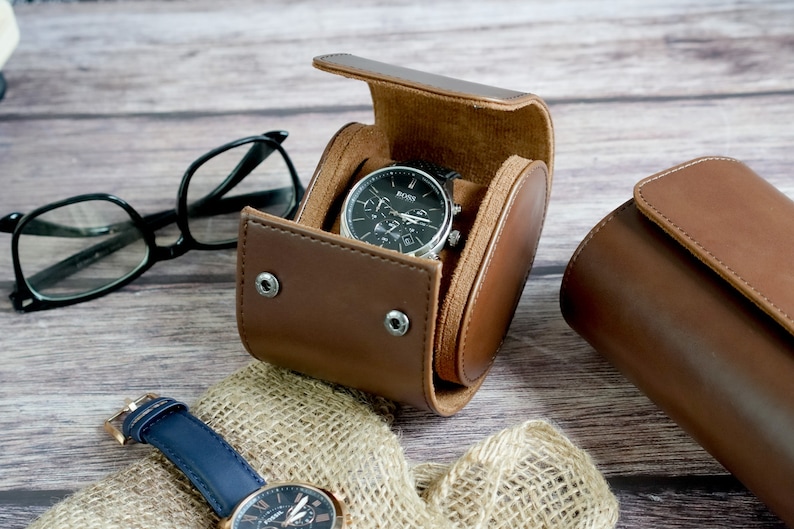 May include: A brown leather watch roll case with a snap button closure containing a black Boss wristwatch with a silver bezel. A second wristwatch with a blue leather strap and a pair of black-rimmed eyeglasses are positioned nearby.