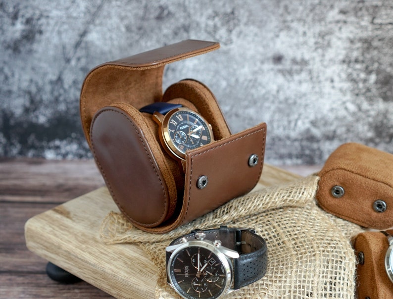 May include: A brown leather watch winder with two watches inside. The winder is open and the watches are visible. The watches have silver and gold accents. The winder is sitting on a wooden surface.