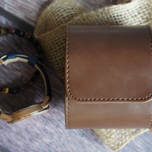 May include: A cylindrical brown leather watch case with visible stitching, positioned next to a wristwatch with a dark blue strap and a beaded bracelet featuring dark brown and black stones, all arranged in a compact display.