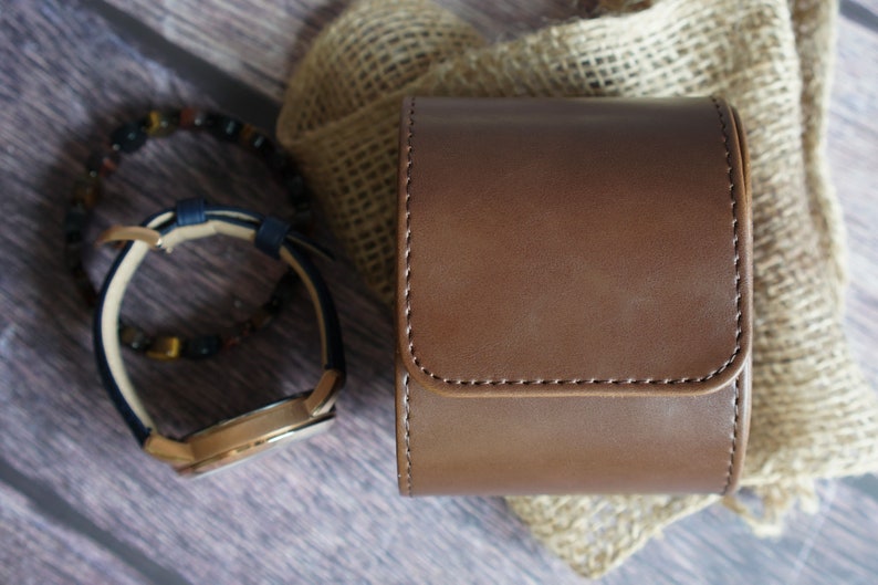 May include: A cylindrical brown leather watch case with visible stitching, positioned next to a wristwatch with a dark blue strap and a beaded bracelet featuring dark brown and black stones, all arranged in a compact display.