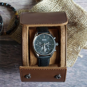 May include: A Hugo Boss chronograph watch with a black perforated leather strap and a dark gray dial, housed in a brown cylindrical leather travel case with a soft tan interior lining and two silver-toned snap button closures on the front flap.