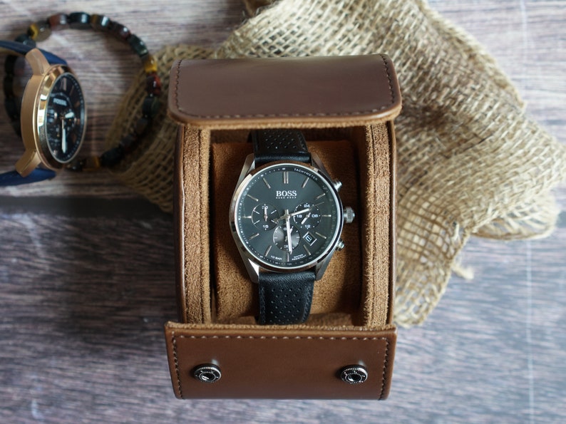 May include: A Hugo Boss chronograph watch with a black perforated leather strap and a dark gray dial, housed in a brown cylindrical leather travel case with a soft tan interior lining and two silver-toned snap button closures on the front flap.