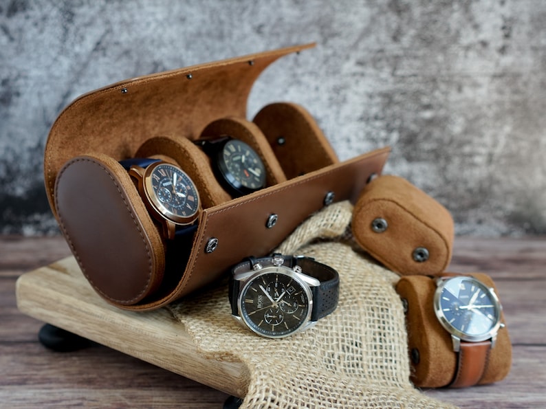 May include: Brown leather watch roll with four slots for watches. The roll is open and three watches are visible. The watches have black, blue, and brown bands. The roll is sitting on a wooden surface.