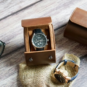 May include: Two leather watch boxes, one open with a silver and black chronograph watch inside, and the other closed. A second watch with a gold face and blue band is on a burlap surface.