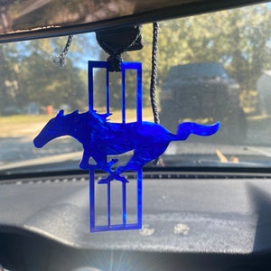 May include: A blue car charm featuring the Ford Mustang logo. The logo is a blue silhouette of a running horse with three vertical bars behind it.